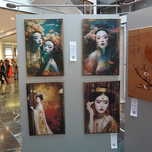 May include: Four framed paintings on display at an art exhibition. The paintings feature women in traditional Asian clothing and imagery. One painting depicts a woman with a crown and jewelry, another shows two women with flowers in their hair, and a third features a woman with a flower in her hair and a fish behind her. The fourth painting is a landscape with a branch of cherry blossoms and a large orange circle.