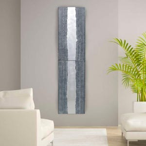 a modern living room with a minimalist design. The focal point is a tall, vertical gray and white artwork hanging on the wall. The room is furnished with two white sofas and a large green plant, creating a clean and elegant atmosphere.