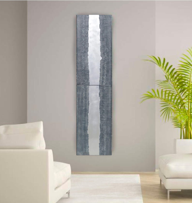 a modern living room with a minimalist design. The focal point is a tall, vertical gray and white artwork hanging on the wall. The room is furnished with two white sofas and a large green plant, creating a clean and elegant atmosphere.