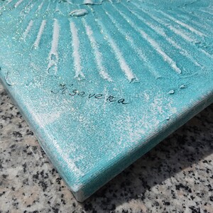 a close-up view of a textured surface, possibly a piece of artwork or a decorative object, with a turquoise or blue-green hue. The surface appears to be made of a material with a textured or embossed appearance, and there is a signature or text written on it.