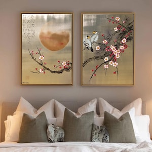 a serene bedroom scene with a large bed adorned with white pillows and a gray comforter. Above the bed, two framed paintings hang on the wall, one depicting a cherry blossom tree with pink flowers and the other showing a bird perched on a branch. The room is illuminated by two pendant lights hanging from the ceiling, creating a warm and inviting atmosphere.