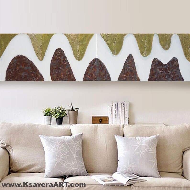 a living room with a beige sofa and two white throw pillows. Above the sofa, there is a large abstract painting with a mix of green, brown, and white colors. On the wall, there are several books and decorative items, including a small wooden box and a potted plant.