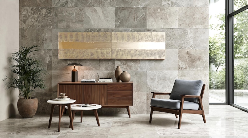 a modern living room with a wooden cabinet, a gray armchair, a round coffee table, and a potted plant. The room has a neutral color scheme with gray and white tiles on the walls and a large window providing natural light.