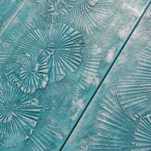 a close-up view of a textured surface with a pattern of overlapping, circular shapes painted in a turquoise color. The surface appears to be made of wood or a similar material, with a rough, textured finish.