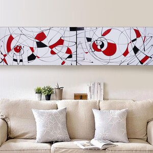 a living room with a beige sofa and a white abstract painting hanging on the wall above it. The painting has a mix of red and black shapes, creating a striking contrast against the white background. The room also includes a few decorative elements, such as potted plants and books, adding to the overall aesthetic.