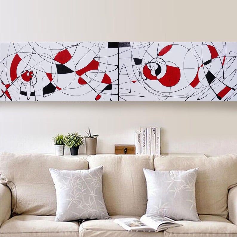 a living room with a beige sofa and a white abstract painting hanging on the wall above it. The painting has a mix of red and black shapes, creating a striking contrast against the white background. The room also includes a few decorative elements, such as potted plants and books, adding to the overall aesthetic.