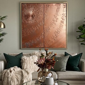 a living room with a large, ornate copper-colored artwork hanging on the wall above a beige sofa adorned with throw pillows. In front of the sofa, there is a glass coffee table with a vase of flowers and a white mug. The room also includes a potted plant and a lamp, creating a cozy and inviting atmosphere.