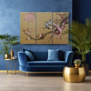 a modern living room with a blue velvet sofa, a gold side table, and a gold coffee table. Above the sofa, there is a large gold-framed painting depicting a bird perched on a branch with pink and white flowers. The room also includes a potted plant and a vase, adding to the overall aesthetic.