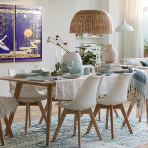Puede incluir: Un conjunto de comedor con mesa de madera clara, cuatro sillas blancas con patas de madera y una silla de piel sint&eacute;tica blanca. La mesa est&aacute; puesta con un camino de mesa blanco, un jarr&oacute;n degradado azul, cristaler&iacute;a y platos bajo una l&aacute;mpara.