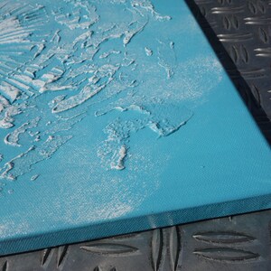a close-up view of a blue canvas with a textured surface, possibly created using a technique like sanding or scratching. The canvas is placed on a metal surface, which has a diamond pattern.