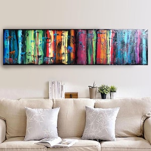 a long, colorful painting hanging on a wall above a beige couch with two white throw pillows. The painting has a vibrant, abstract design with vertical stripes in various colors. Below the painting, there are some books and potted plants on a shelf, creating a cozy and inviting atmosphere in the living room.