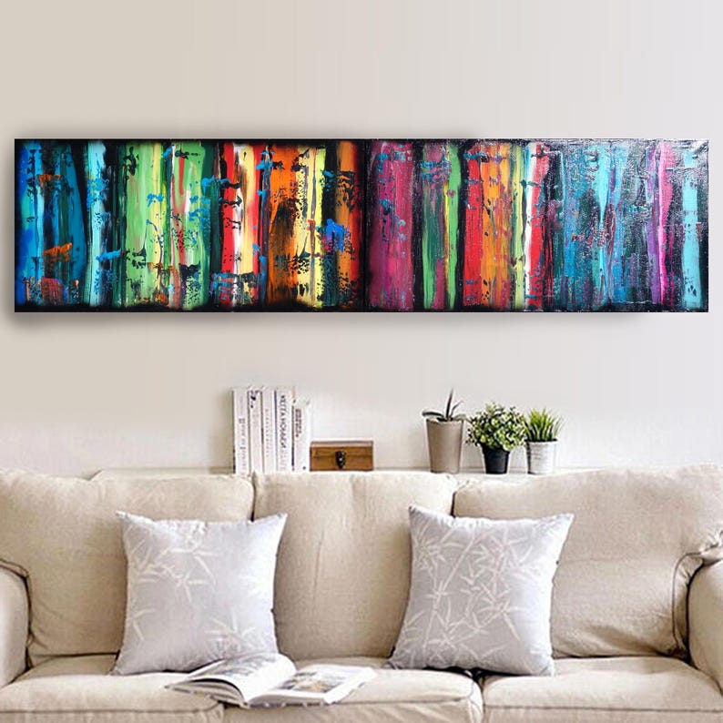 a long, colorful painting hanging on a wall above a beige couch with two white throw pillows. The painting has a vibrant, abstract design with vertical stripes in various colors. Below the painting, there are some books and potted plants on a shelf, creating a cozy and inviting atmosphere in the living room.