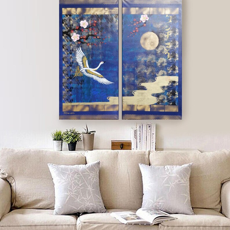 a cozy living room with a beige sofa and a bookshelf below it. Above the sofa, there is a large painting depicting a white crane in flight over a blue landscape with a full moon and cherry blossoms.