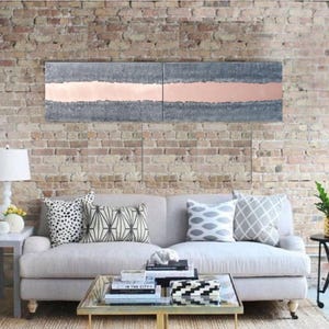 a modern living room with a gray sofa adorned with patterned throw pillows, a glass coffee table, and a brick wall backdrop. Above the sofa, there is a large abstract painting with a horizontal stripe in shades of gray and pink.