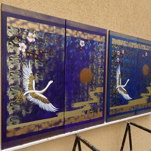 two large paintings hanging on a wall, depicting a crane in flight against a backdrop of purple and gold hues.
