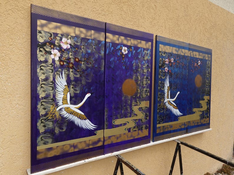 two large paintings hanging on a wall, depicting a crane in flight against a backdrop of purple and gold hues.