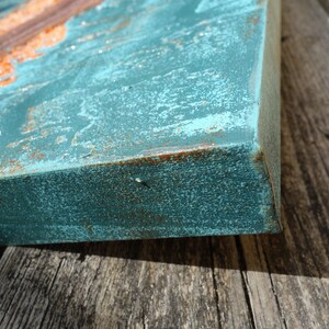 a close-up view of a weathered, blue-green surface with a rust-colored element, possibly a metal or metal-like material, attached to it. The surface appears to be a wooden board or piece of wood, with a rough, textured appearance.