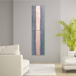 a modern living room with a tall vertical artwork hanging on the wall. The artwork has a pink stripe running through it, creating a striking visual contrast against the gray wall. The room is furnished with two white sofas, and a large potted plant is placed on the right side of the room, adding a touch of greenery to the space.
