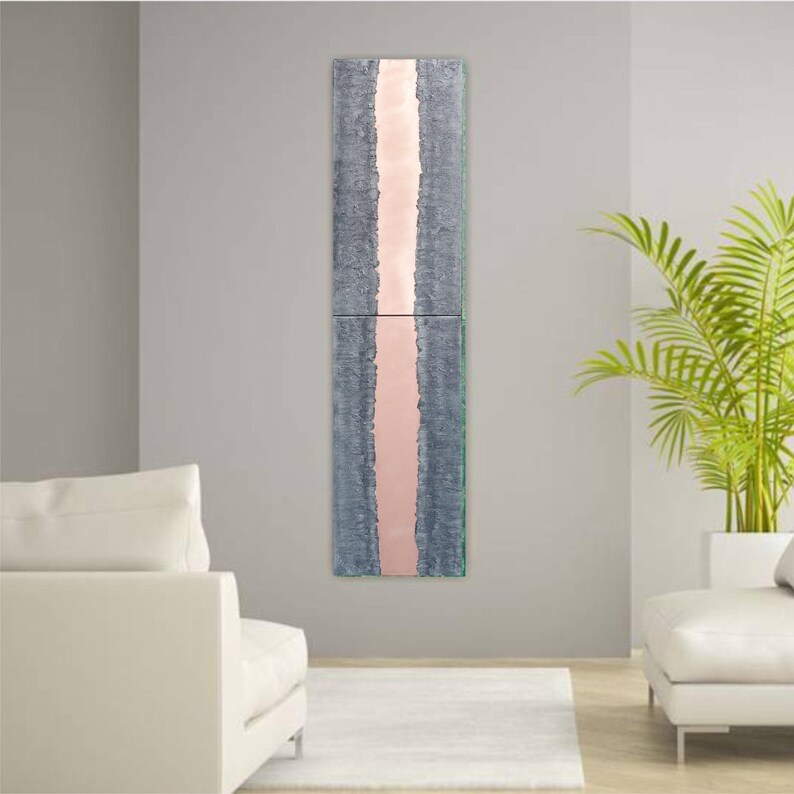 a modern living room with a tall vertical artwork hanging on the wall. The artwork has a pink stripe running through it, creating a striking visual contrast against the gray wall. The room is furnished with two white sofas, and a large potted plant is placed on the right side of the room, adding a touch of greenery to the space.