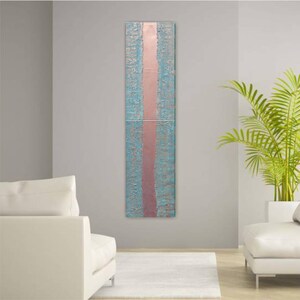 a modern living room with a tall vertical artwork hanging on the wall. The artwork has a blue and pink color scheme and appears to be a painting or a decorative piece. The room is furnished with two white sofas and a large potted plant, creating a clean and minimalist aesthetic.