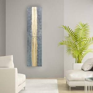 a modern living room with a gray and gold vertical artwork hanging on the wall. The room is furnished with two white sofas and a large potted plant, creating a serene and stylish atmosphere.