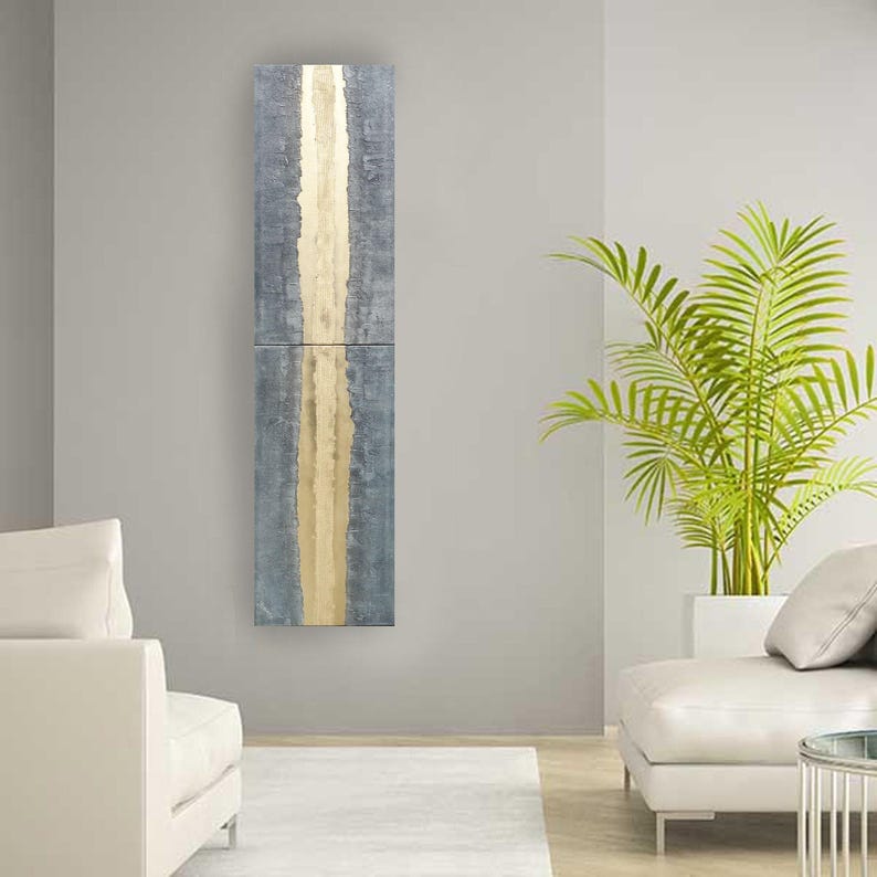 a modern living room with a gray and gold vertical artwork hanging on the wall. The room is furnished with two white sofas and a large potted plant, creating a serene and stylish atmosphere.