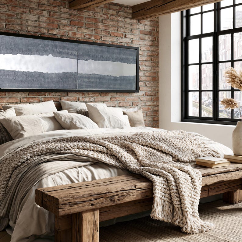 a cozy bedroom with a large bed, a wooden bench, and a brick wall with a large window. The bed is covered in a white comforter and has a throw blanket draped over it. The wooden bench is positioned next to the bed, and a vase is visible on the windowsill. The brick wall adds a rustic touch to the room, and a large window allows natural light to enter the space.