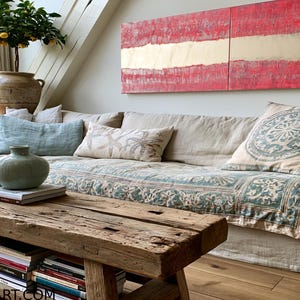 a cozy living room with a large, comfortable couch adorned with patterned pillows and a throw blanket. A wooden coffee table sits in front of the couch, and a large, colorful abstract painting hangs on the wall above it. The room is decorated with various plants and books, creating a warm and inviting atmosphere.