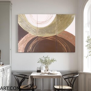 a modern kitchen with a large abstract painting hanging on the wall above a wooden dining table with two chairs. The kitchen is well-decorated with various plants and decorative items, creating a cozy and inviting atmosphere.