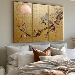 a large, gold-colored painting of a bird perched on a branch with flowers, hanging above a bed with white pillows and a brown throw pillow.