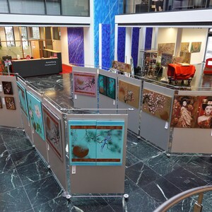 an art exhibition featuring various framed artworks displayed on walls and pedestals throughout a spacious room with marble floors and large windows.