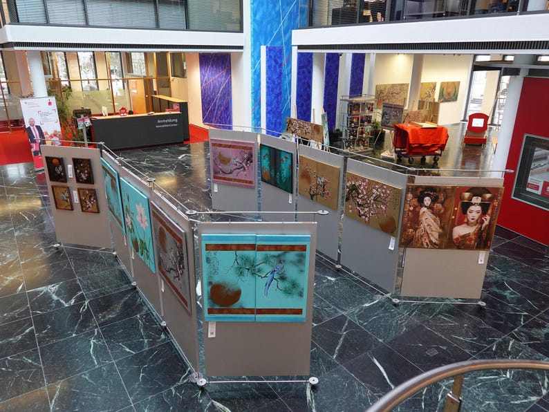 an art exhibition featuring various framed artworks displayed on walls and pedestals throughout a spacious room with marble floors and large windows.