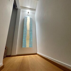 a hallway with a wooden floor and white walls. A large blue and white abstract artwork is hanging on the wall, creating a focal point in the space.