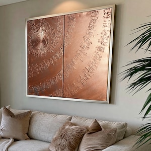 a large, square, metallic artwork with a textured, metallic surface hanging on a wall above a beige couch with plush pillows.