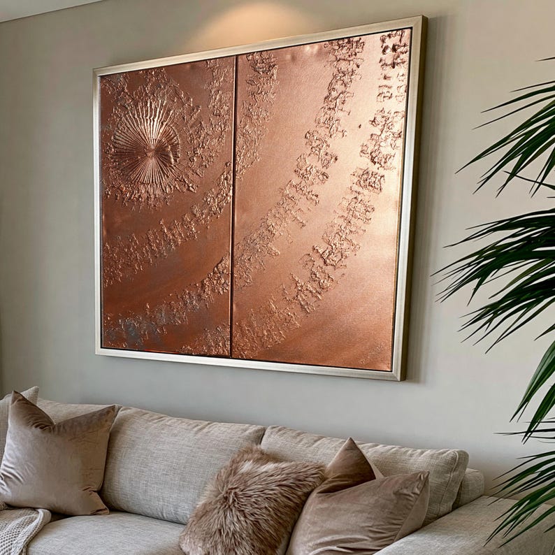 a large, square, metallic artwork with a textured, metallic surface hanging on a wall above a beige couch with plush pillows.