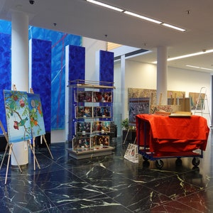 Puede incluir: Interior de una galer&iacute;a de arte con pinturas sobre caballetes, grabados enmarcados en una vitrina y grandes obras de arte murales azules abstractas. Un carro rojo est&aacute; cubierto con una tela roja.