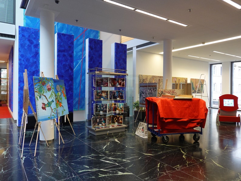 Puede incluir: Interior de una galer&iacute;a de arte con pinturas sobre caballetes, grabados enmarcados en una vitrina y grandes obras de arte murales azules abstractas. Un carro rojo est&aacute; cubierto con una tela roja.