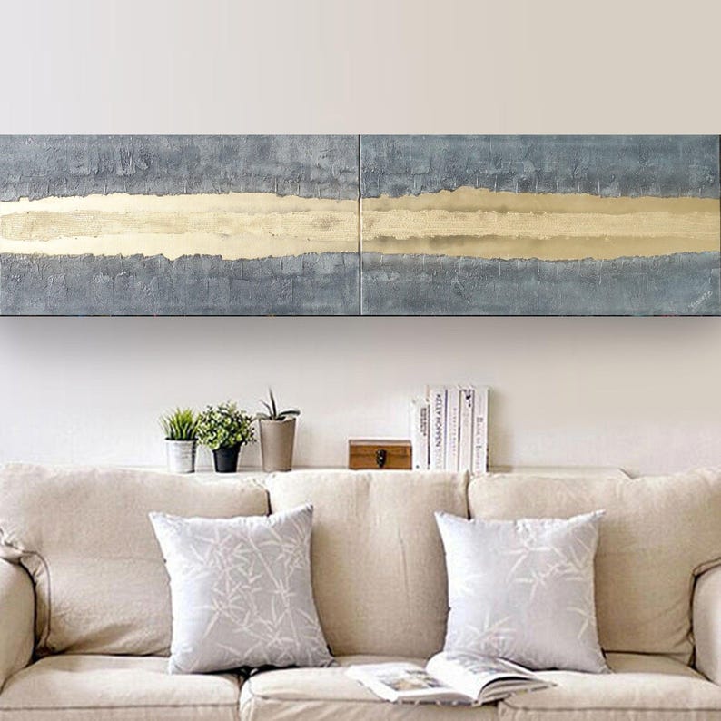 a cozy living room with a beige sofa adorned with two patterned throw pillows. Above the sofa, there is a large abstract painting with a horizontal stripe of blue and gold colors. On the wall, there is a shelf holding several books and a small wooden box.