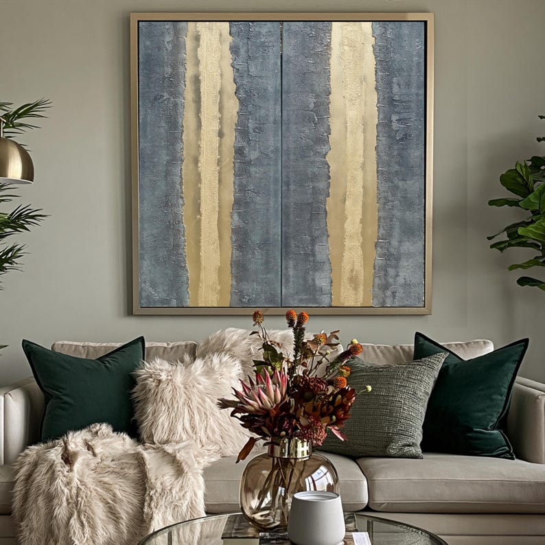 a cozy living room with a large abstract painting featuring vertical lines in shades of blue and gold hanging on the wall above a beige sofa adorned with green and gray throw pillows. In front of the sofa, there is a glass coffee table with a vase of flowers and a white mug.