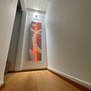 a hallway with a wooden floor and a large abstract painting hanging on the wall. The painting features a mix of orange and brown colors, creating a vibrant and dynamic visual effect. The hallway appears to be well-lit, with a light source visible in the top left corner.