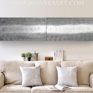 a cozy living room with a beige sofa adorned with two gray throw pillows. Above the sofa, there is a large, textured metal panel that adds a modern touch to the space. On the shelf below the sofa, there are several books and decorative items, creating a welcoming atmosphere.