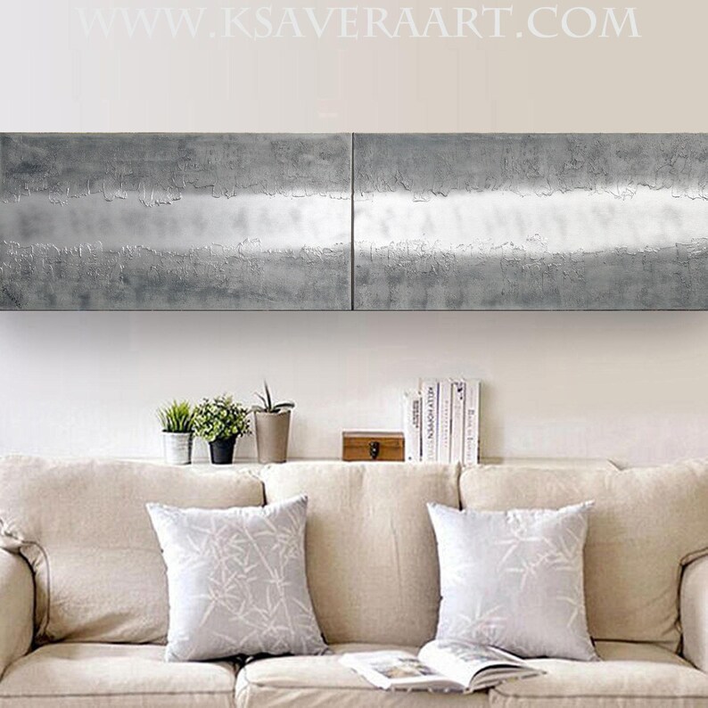 a cozy living room with a beige sofa adorned with two gray throw pillows. Above the sofa, there is a large, textured metal panel that adds a modern touch to the space. On the shelf below the sofa, there are several books and decorative items, creating a welcoming atmosphere.