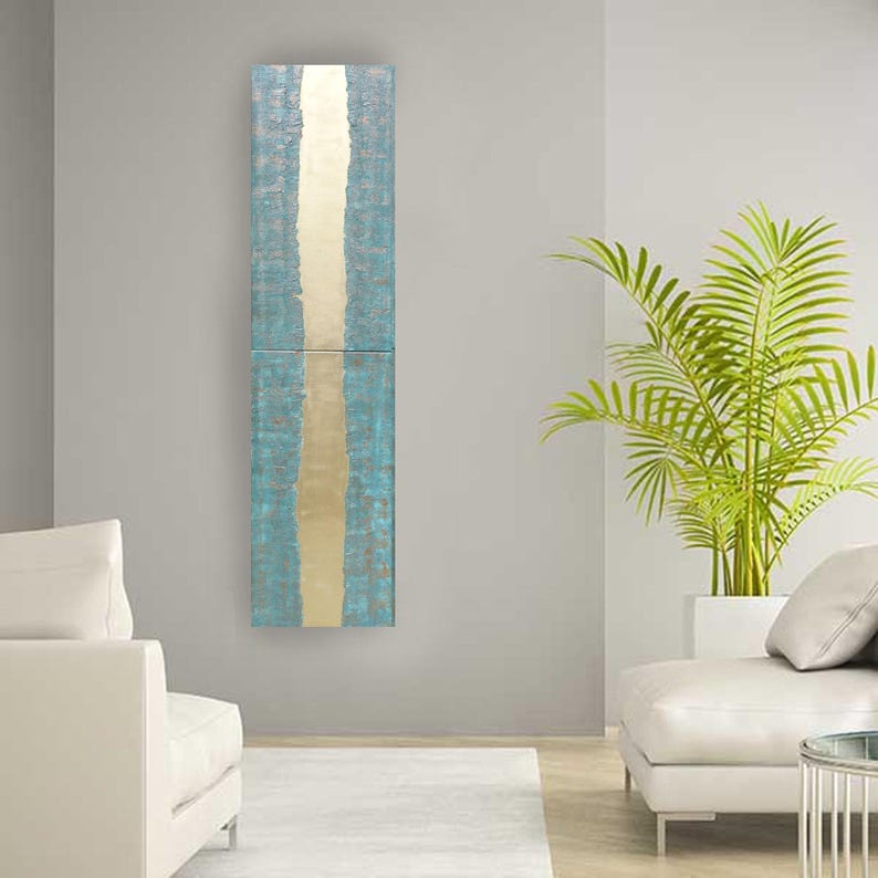 a modern living room with a tall, vertical blue and gold artwork hanging on the gray wall. The room is furnished with two white sofas and a large potted plant, creating a serene and stylish atmosphere.