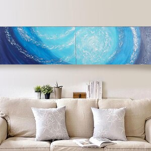 a cozy living room with a beige sofa and a bookshelf filled with books. Above the sofa, there is a large painting depicting a blue and white abstract design, adding a touch of artistic flair to the space.