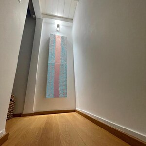 a hallway with a wooden floor and white walls. There is a large abstract painting hanging on the wall, which appears to be a colorful and textured piece. The hallway has a minimalist and clean design, with a single light source illuminating the space.