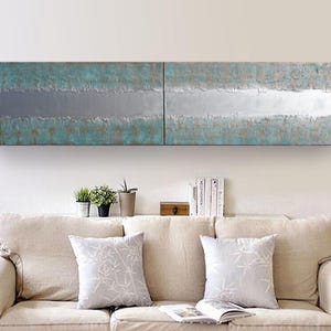 a cozy living room with a beige sofa adorned with two patterned throw pillows. Above the sofa, there is a large, abstract painting with a blue and white color scheme. On the wall, there is a shelf holding several books and a small wooden box.