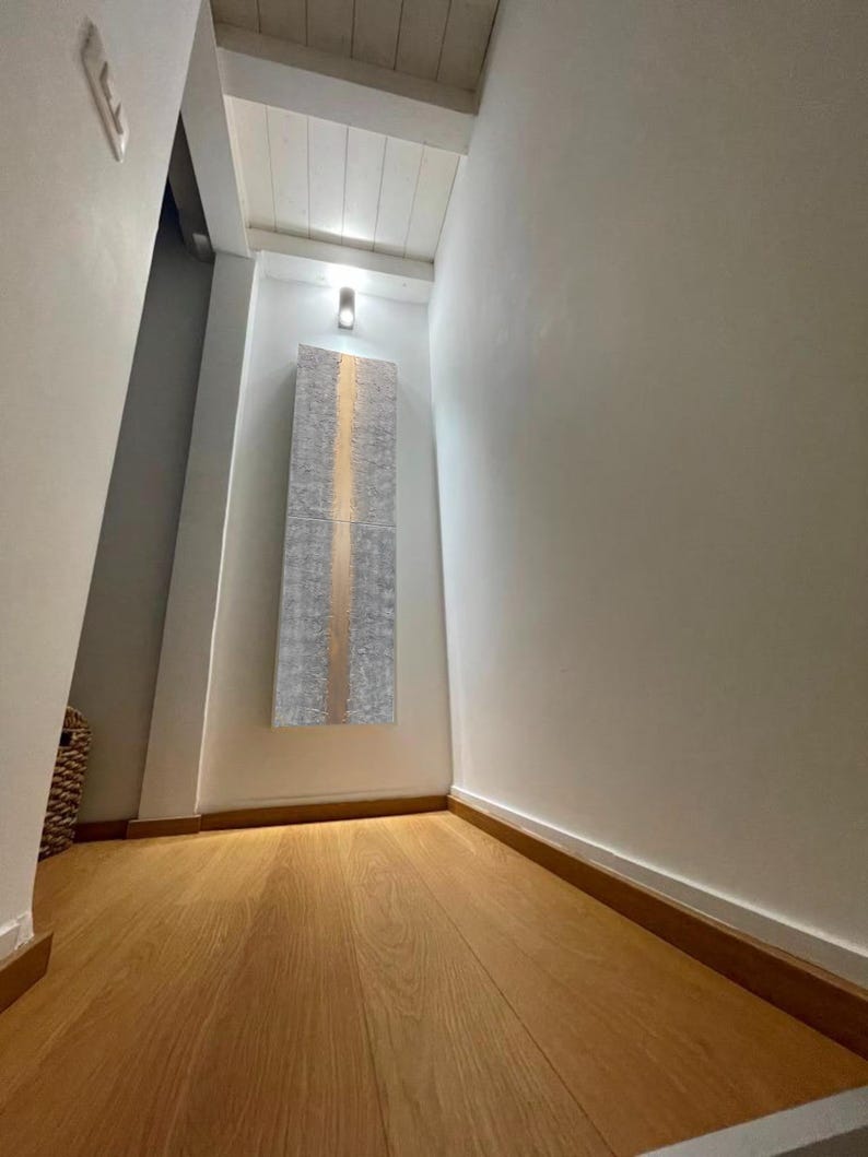 a hallway with a wooden floor and white walls. There is a large piece of artwork hanging on the wall, and a light fixture is visible above it.