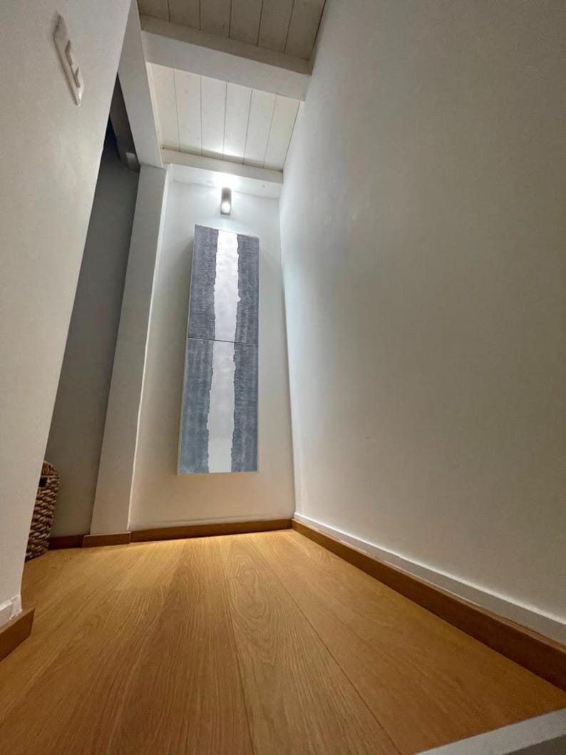 a hallway with a wooden floor and white walls. There is a large piece of artwork hanging on the wall, and a light fixture is visible above it. The hallway appears to be empty and well-lit, with a sense of spaciousness.