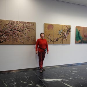 A woman in a red sweater stands in front of three large paintings on a white wall.