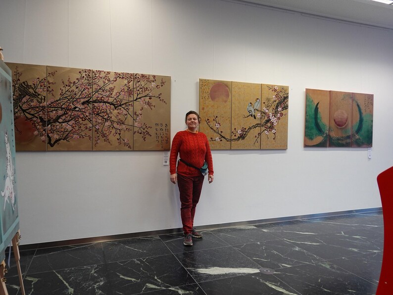 A woman in a red sweater stands in front of three large paintings on a white wall.